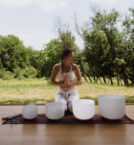 Femme assise en posture de prière devant des bols chantants en cristal de quartz, lors d’une séance de thérapie sonore en pleine nature. Une scène lumineuse qui évoque calme, ancrage et harmonie vibratoire.